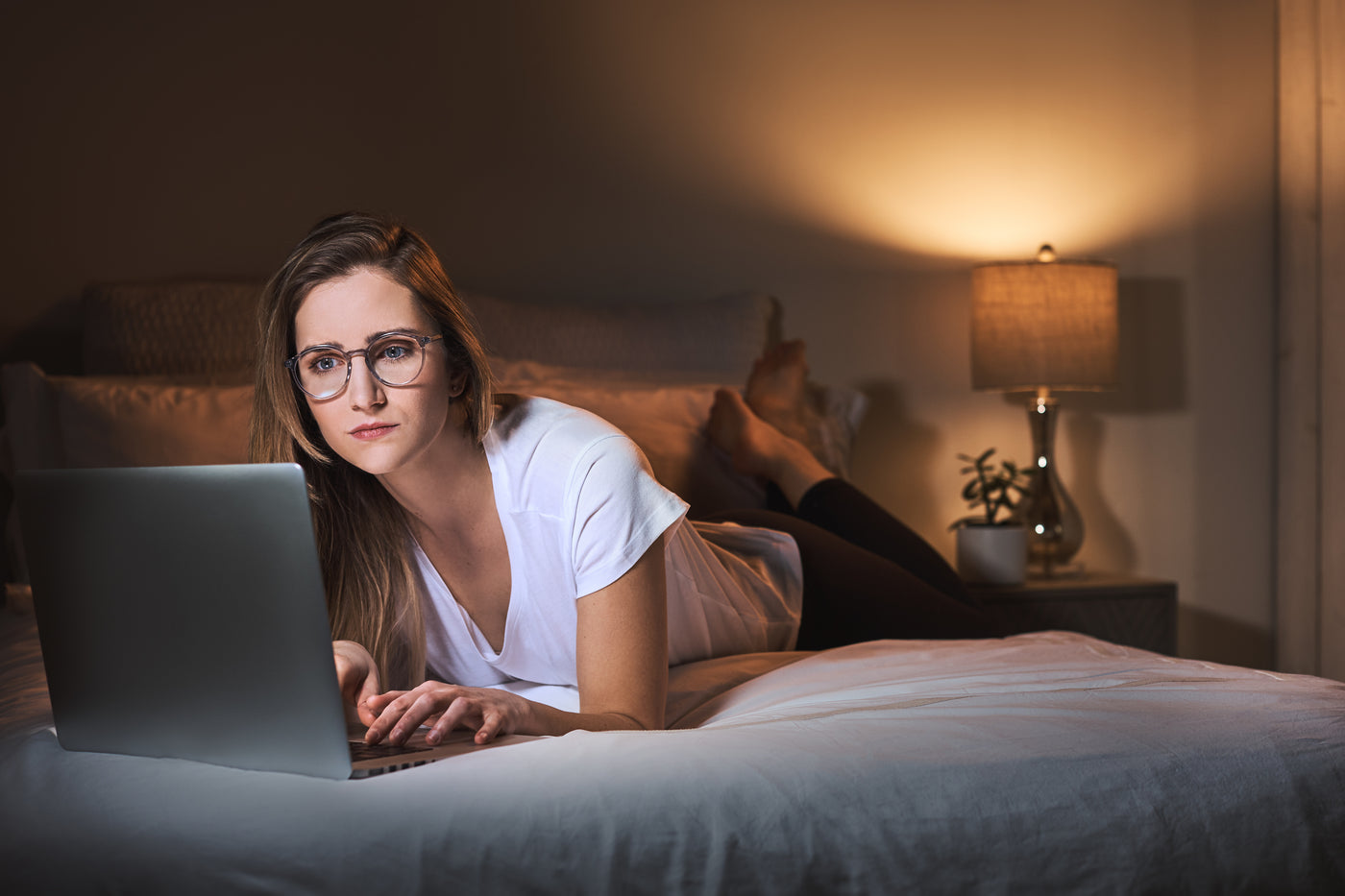 Woman laying on a bed using a laptop and wearing blue light blocking glasses.